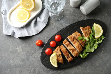 Tasty cut schnitzel served with tomatoes, greens and lemon on grey textured table, flat lay