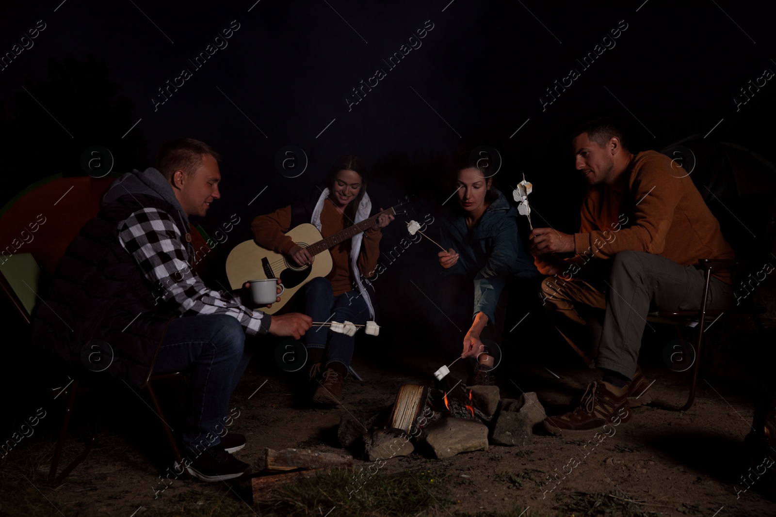 Photo of Group of friends roasting marshmallows on bonfire at camping site in evening