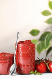 Jars of tasty tomato paste, spoon and ingredients on white table
