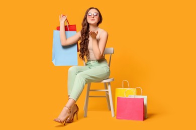Woman holding colorful shopping bags and blowing kiss on chair against orange background