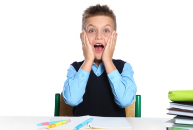 Emotional little boy in uniform with school stationery at desk against white background