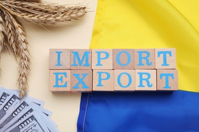 Photo of Words Import and Export made of wooden cubes, ears of wheat, money and Ukrainian flag on beige background, flat lay