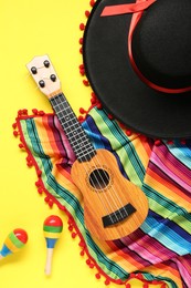 Photo of Mexican sombrero hat, guitar, maracas and colorful poncho on yellow background, flat lay