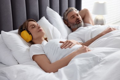 Photo of Smiling woman with headphones lying near her snoring husband in bed at home