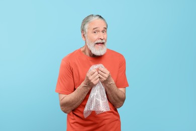 Emotional senior man popping bubble wrap on light blue background. Stress relief