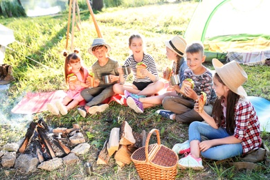 Photo of Little children eating sandwiches near bonfire and tent. Summer camp