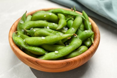 Photo of Bowl of green edamame beans in pods with salt on light grey table, closeup