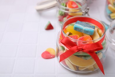 Photo of Glass jars with tasty colorful jelly candies on white tiled table. Space for text