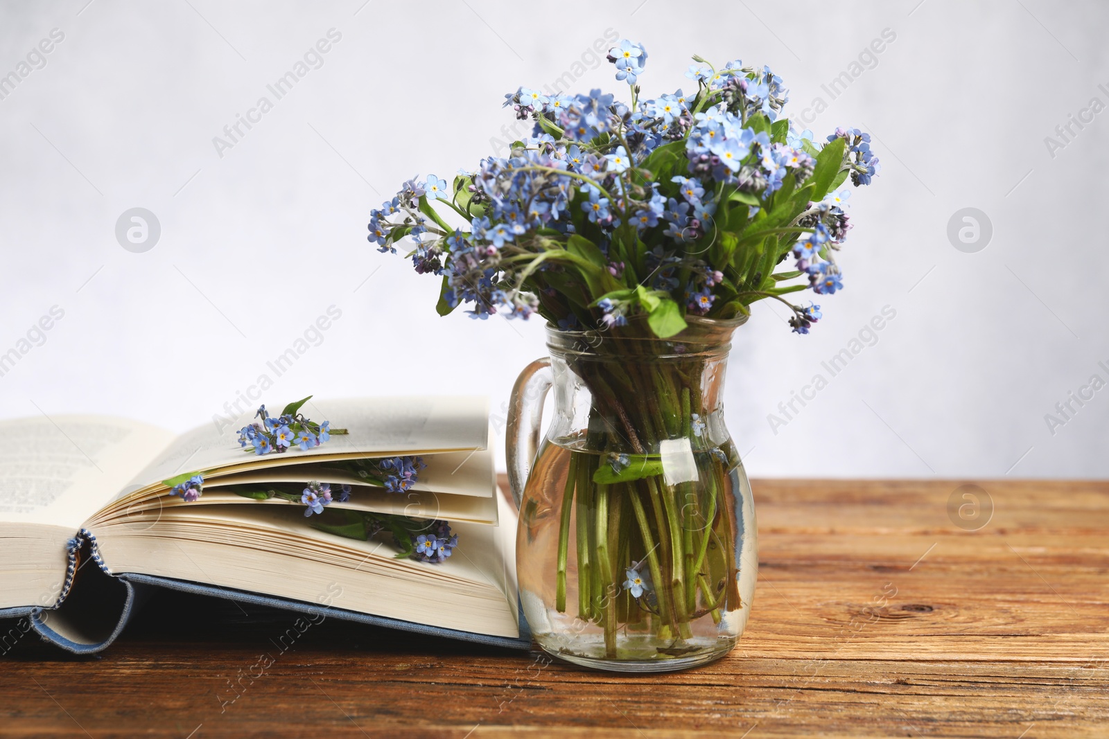 Photo of Bouquet of beautiful forget-me-not flowers in glass jug and book on wooden table against light background