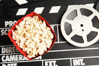 Photo of Tasty popcorn in paper package and film reel on clapperboard, top view. Cinema snack