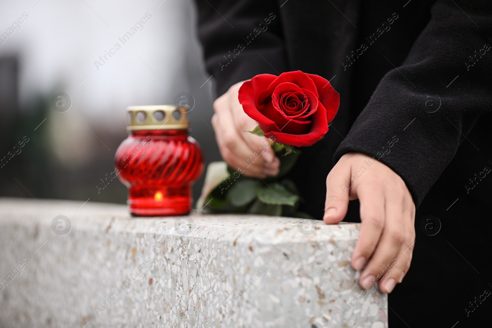 Photo of Woman holding red rose near grey granite tombstone with candle outdoors, closeup. Funeral ceremony