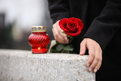 Woman holding red rose near grey granite tombstone with candle outdoors, closeup. Funeral ceremony