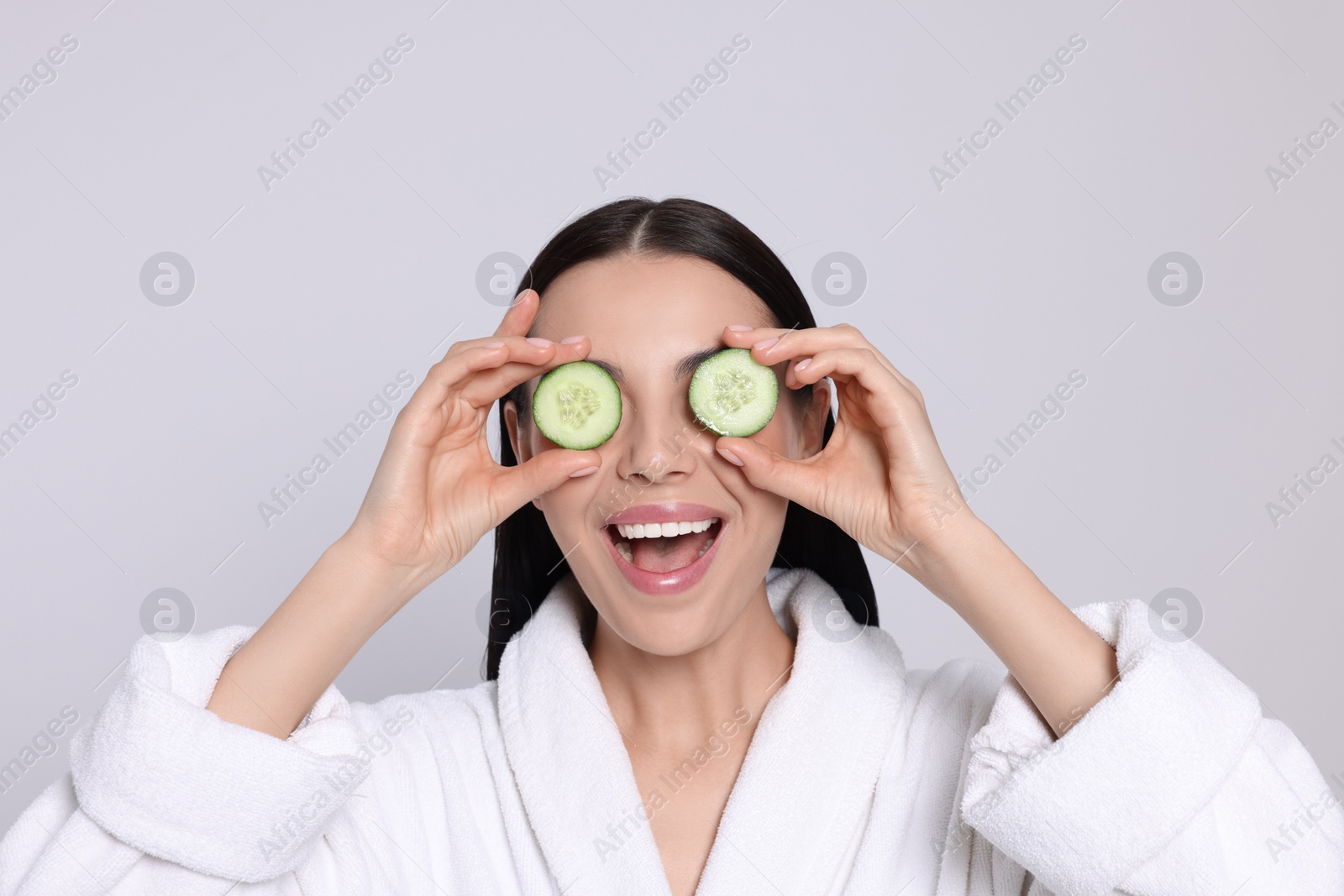 Photo of Woman in bathrobe holding pieces of cucumber on light grey background. Spa treatment