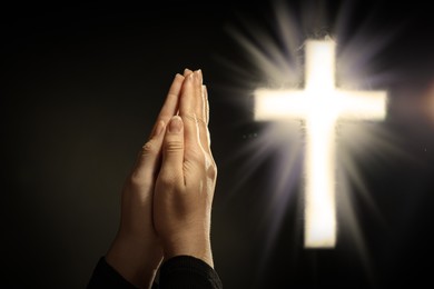 Image of Christian cross and woman holding hands clasped while praying against darkness, closeup