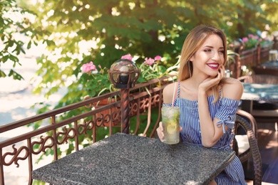 Young woman with glass of tasty lemonade at table in cafe, outdoors. Natural detox drink