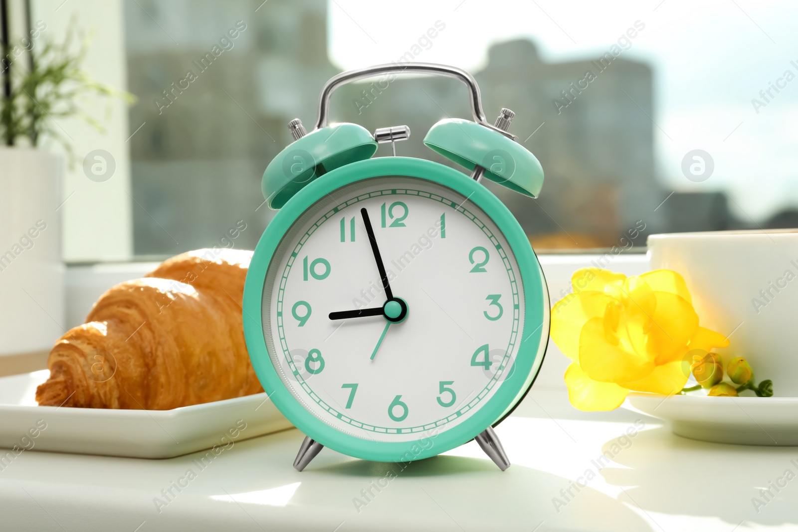 Photo of Alarm clock, croissant and cup of drink with flower on windowsill. Good morning