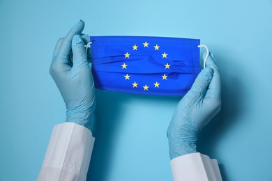 Image of Doctor holding medical mask with European Union flag on light blue background, closeup. Coronavirus outbreak in Europe
