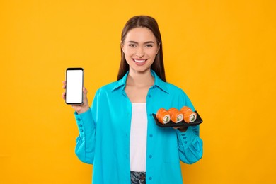 Happy young woman with plate of sushi rolls and smartphone on orange background. Space for text