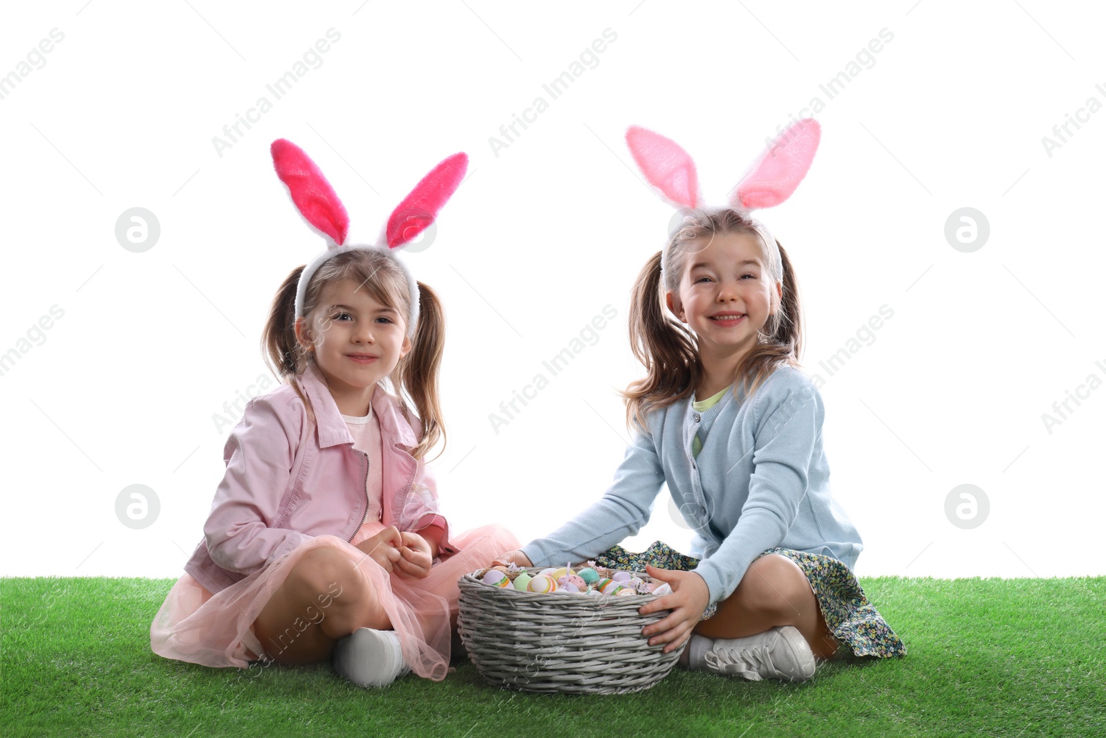 Photo of Adorable little girls with bunny ears and wicker basket full of Easter eggs on green grass against white background