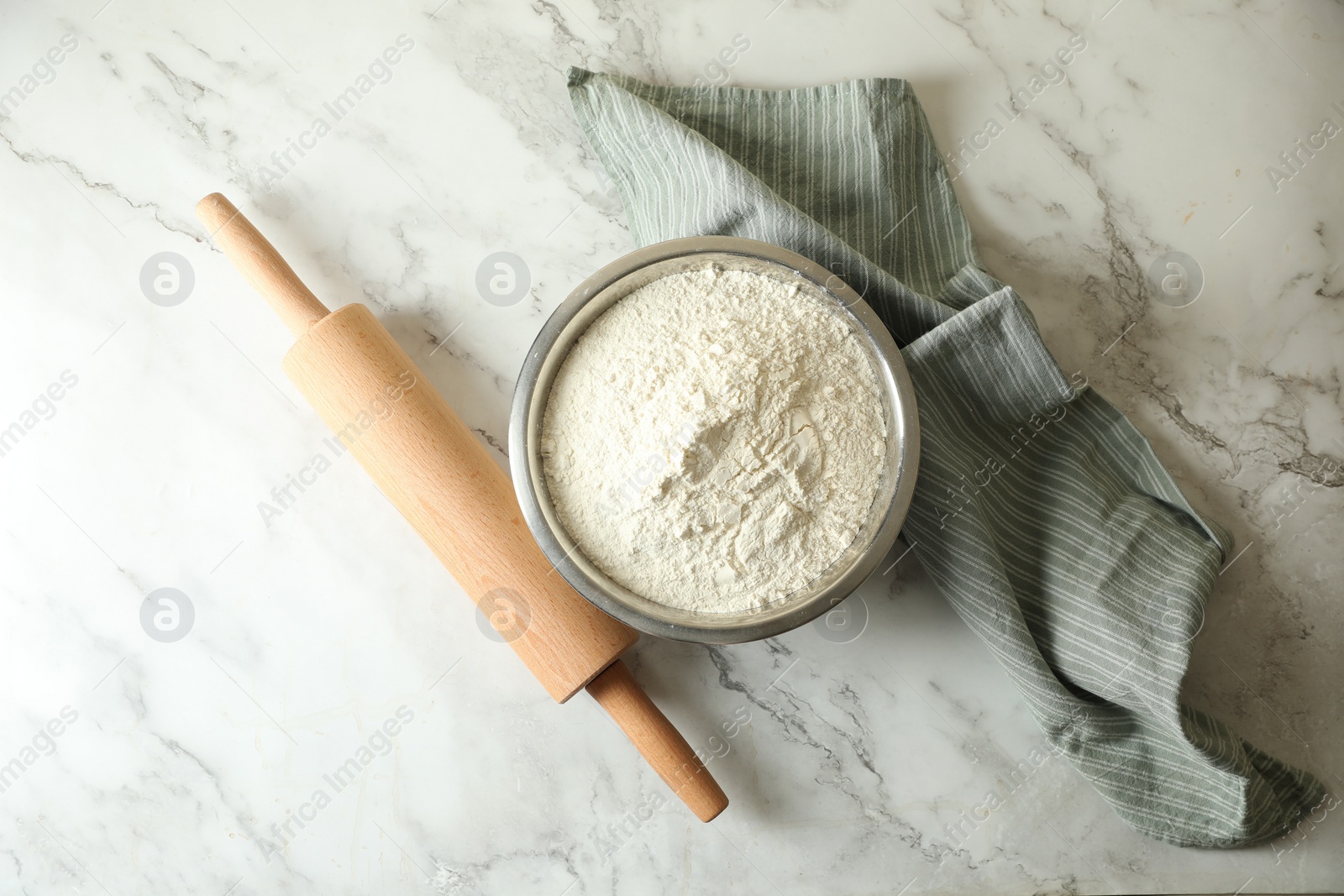 Photo of Flour in bowl, rolling pin and napkin on white marble table, top view