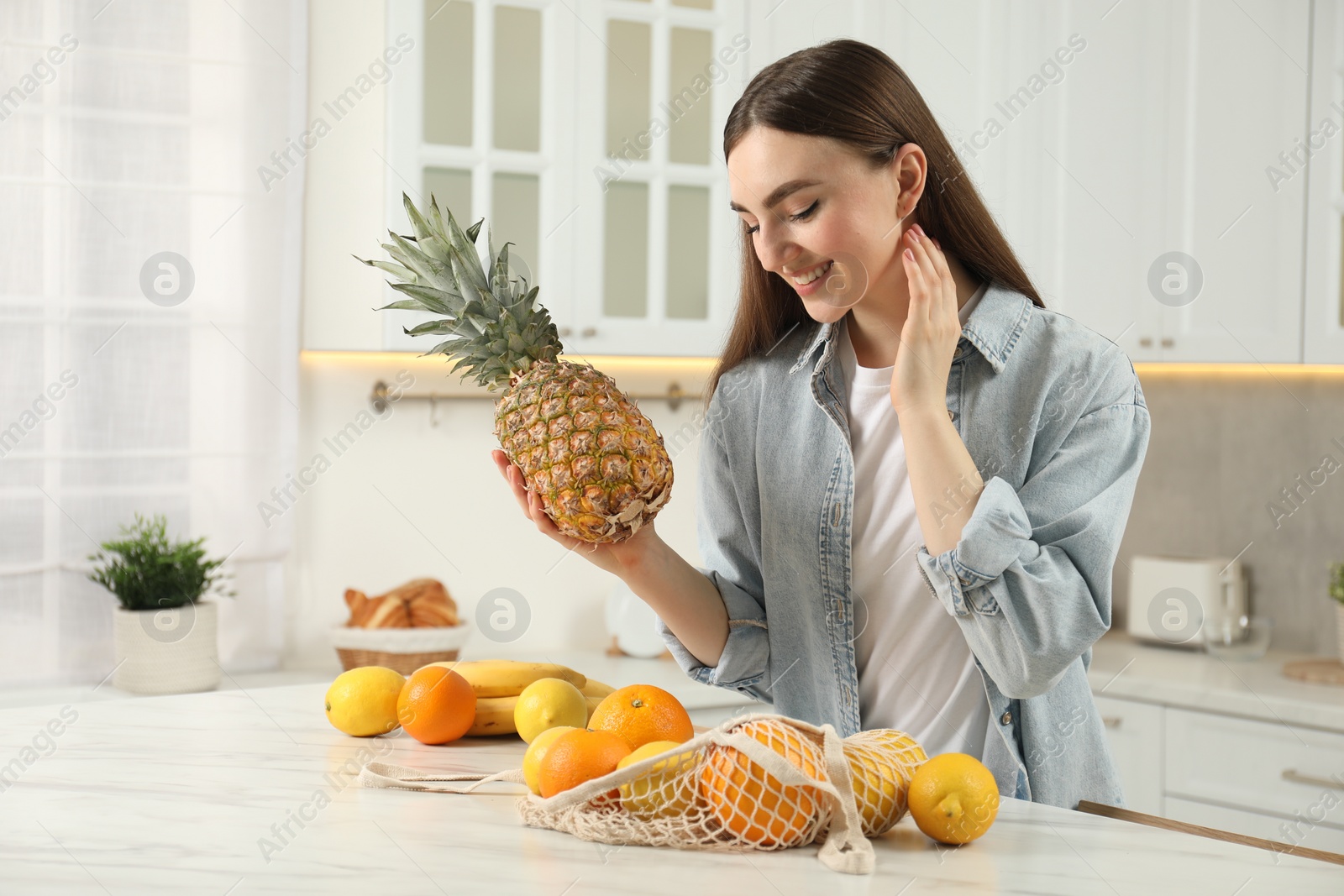 Photo of Woman with pineapple and string bag of fresh fruits at light marble table in kitchen