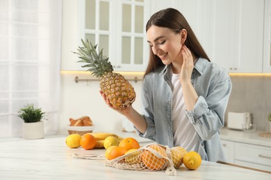 Woman with pineapple and string bag of fresh fruits at light marble table in kitchen
