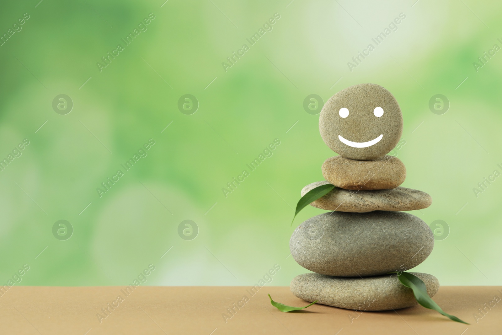 Photo of Stack of stones with drawn happy face and green leaves on table against blurred background, space for text. Zen concept