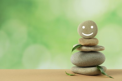 Photo of Stack of stones with drawn happy face and green leaves on table against blurred background, space for text. Zen concept