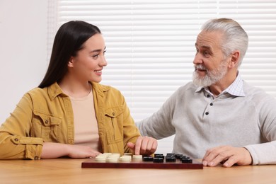 Photo of Playing checkers. Senior man learning young woman at table in room