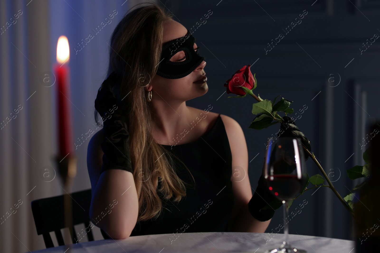 Photo of Elegant woman in black eye mask with rose at table indoors in evening