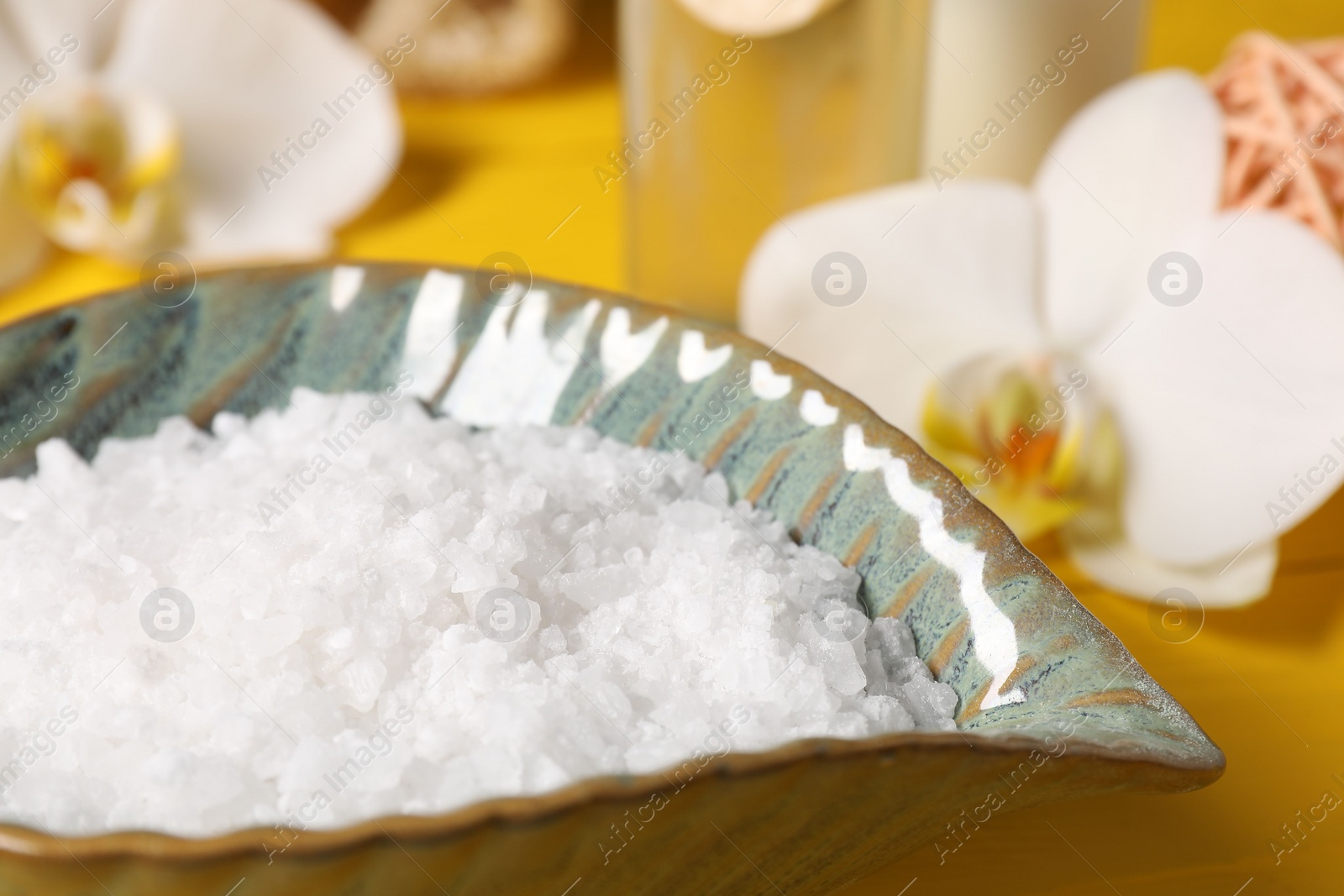 Photo of Natural sea salt in bowl and orchid flowers on yellow table, closeup