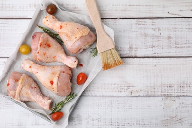 Photo of Raw marinated chicken drumsticks, rosemary, tomatoes and basting brush on white wooden table, flat lay. Space for text