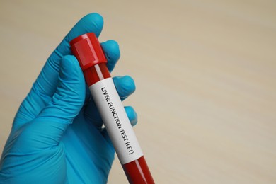Laboratory worker holding tube with blood sample and label Liver Function Test at beige table, closeup. Space for text