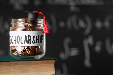 Scholarship concept. Glass jar with coins, graduation cap and book against blackboard, space for text