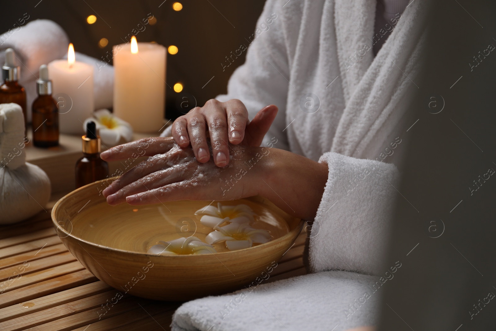 Photo of Woman applying scrub onto her hands in spa, closeup. Bowl of water and flowers on wooden table