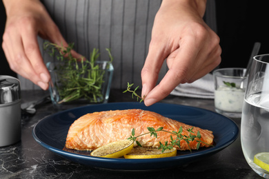 Photo of Woman adding thyme to cooked red fish on dark marble table, closeup