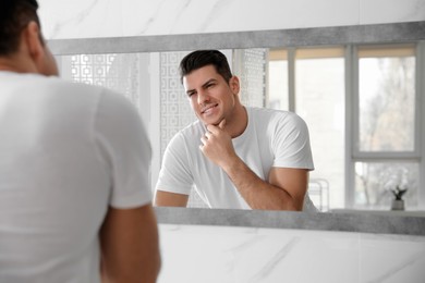 Handsome man touching his smooth face after shaving near mirror in bathroom