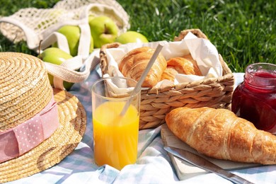 Photo of Blanket with juice, jam and croissants for picnic on green grass, closeup