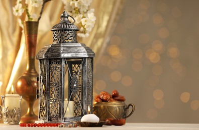 Photo of Arabic lantern, misbaha, candles, dates and flowers on table against blurred lights, space for text
