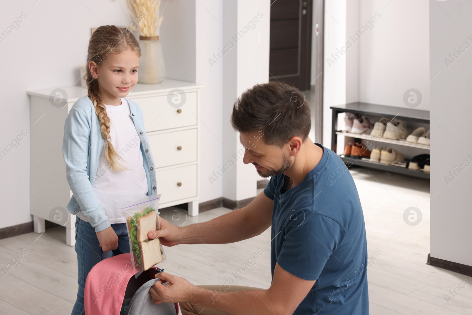 Photo of Happy father putting lunch box into daughter`s backpack at home. Preparing to school