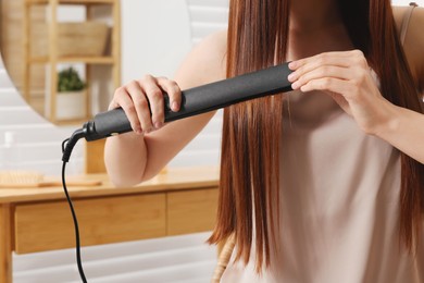 Photo of Young woman using hair iron in room, closeup
