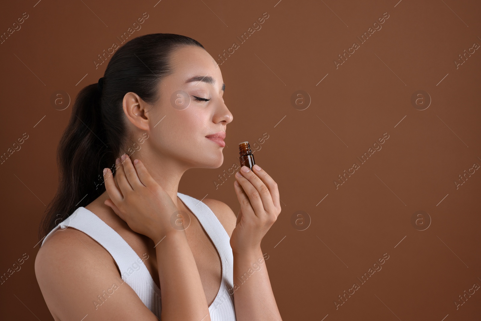 Photo of Beautiful young woman with bottle of essential oil on brown background, space for text