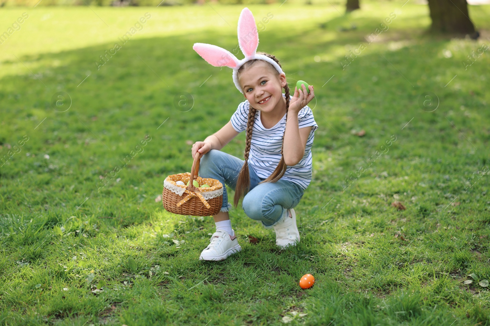 Photo of Easter celebration. Cute little girl in bunny ears hunting eggs outdoors