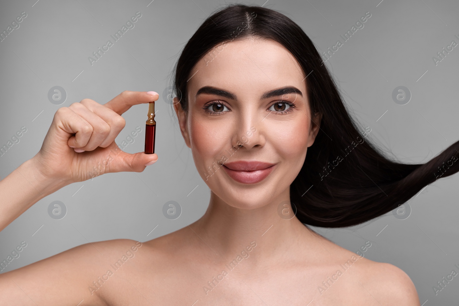 Photo of Beautiful young woman with long healthy hair holding ampoule on grey background