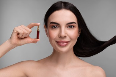 Beautiful young woman with long healthy hair holding ampoule on grey background