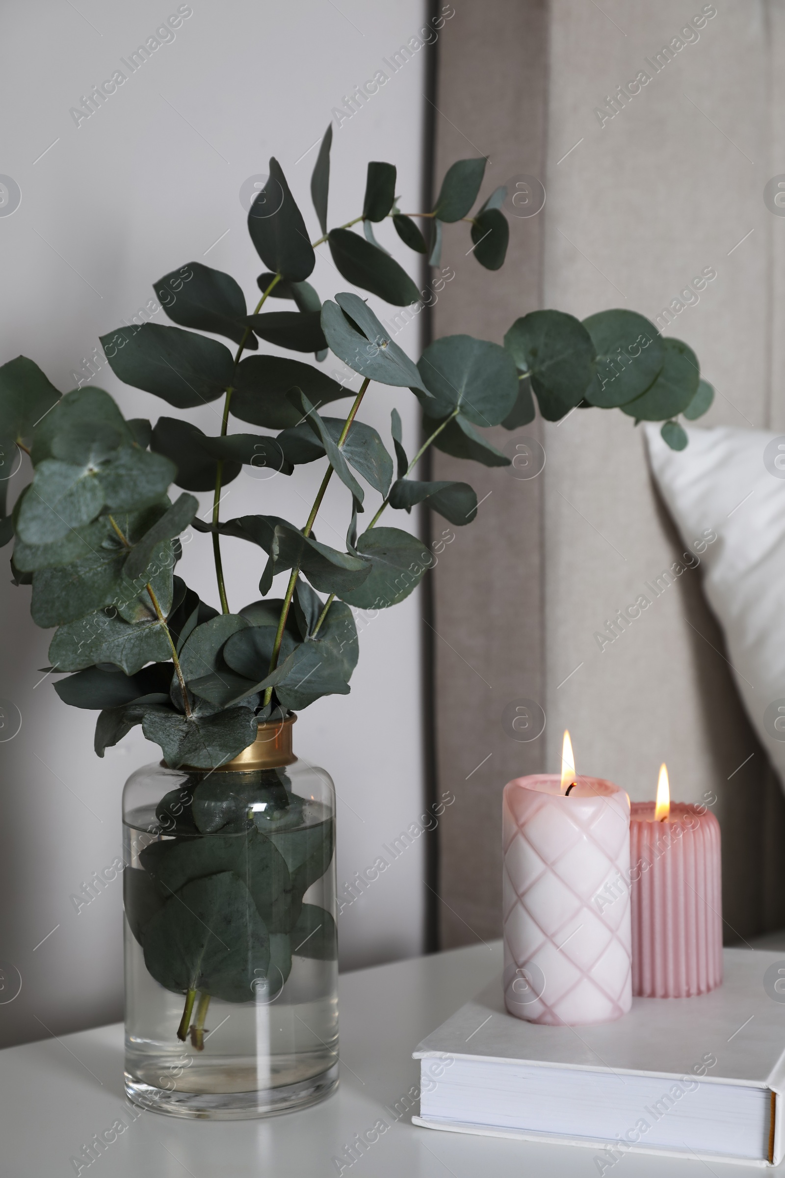 Photo of Vase with beautiful eucalyptus branches, book and candles on nightstand in bedroom