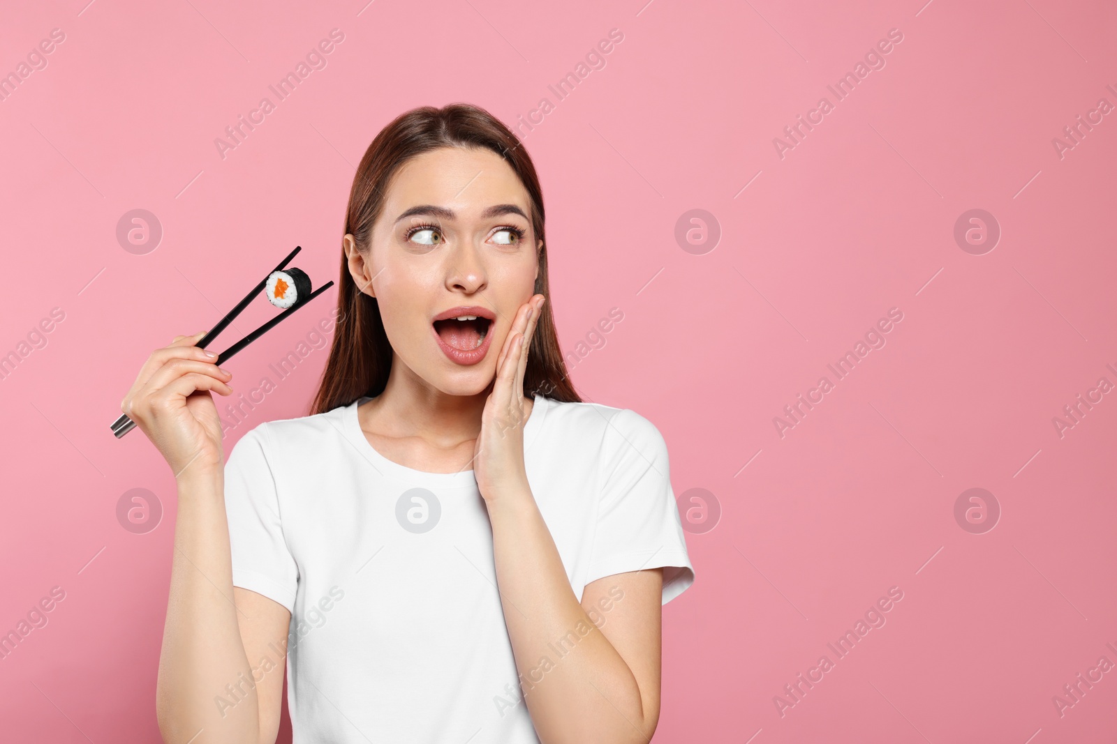 Photo of Emotional young woman holding sushi roll with chopsticks on pink background. Space for text