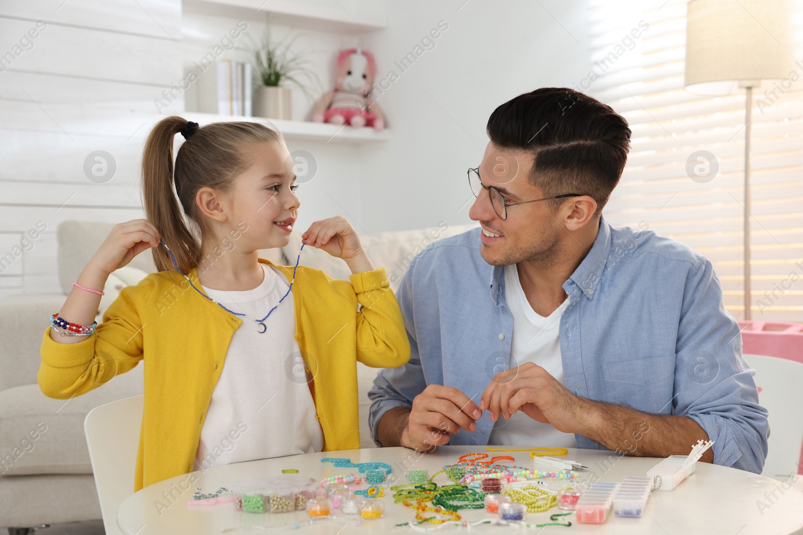 Photo of Happy father with his cute daughter making beaded jewelry at table in room