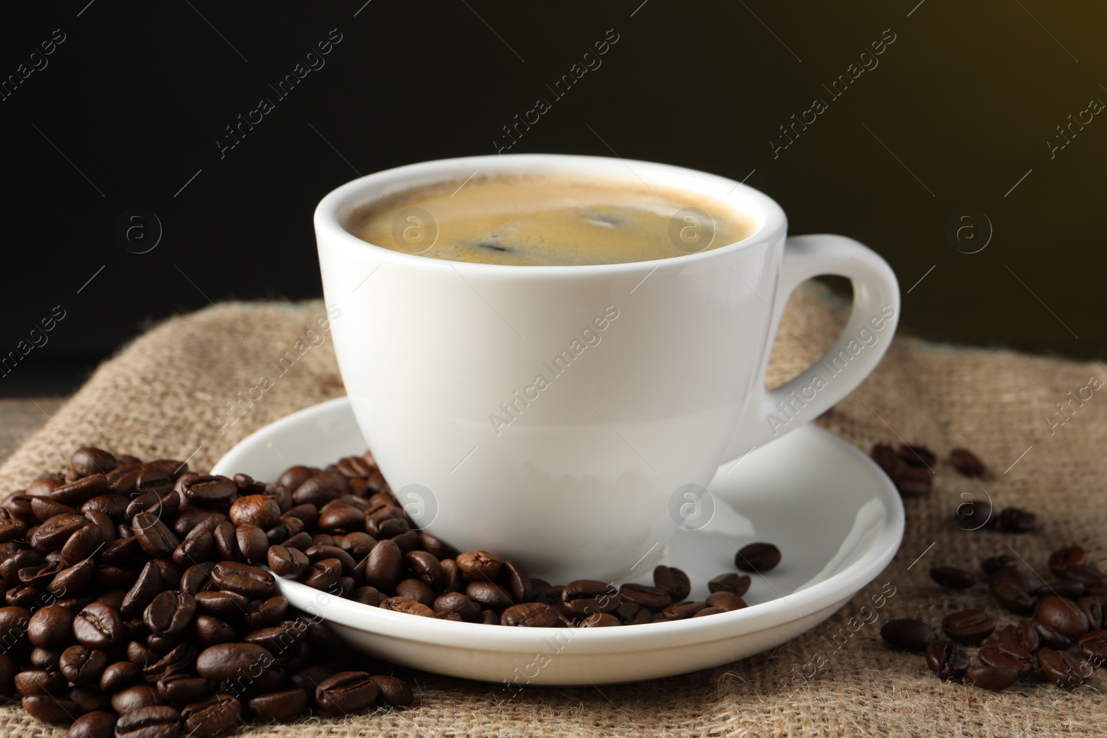 Photo of Cup of hot aromatic coffee and roasted beans on sackcloth against dark background