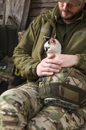 Photo of Ukrainian soldier in uniform warming little stray cat indoors, closeup